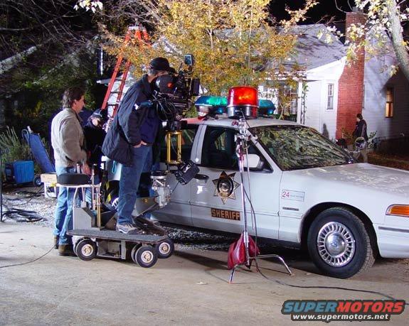 Sheriff's car with lights on, film crew around it, house in background