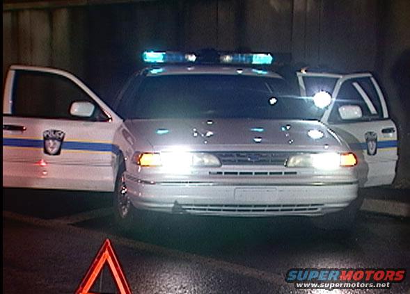 White 1997 Ford Crown Victoria police car with doors open, emergency lights on, and a reflective triangle on the road