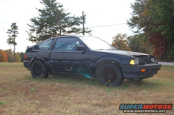 Black hatchback with missing front grille and hood open, parked on dry grass
