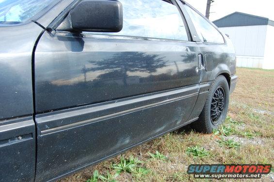 Side view of a dark-colored car's door and rear wheel with a mesh-style rim