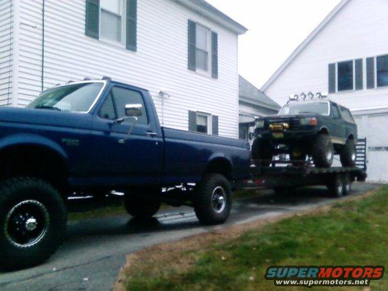 Dark green Ford Bronco on a flatbed trailer, lifted with large tires and a winch. Towed by a blue Ford pickup truck