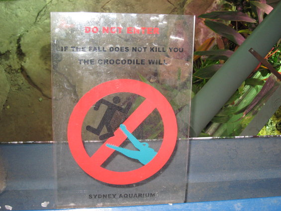 Sign with red prohibition circle over a running person and a crocodile, text reads 'DO NOT ENTER IF THE FALL DOES NOT KILL YOU THE CROCODILE WILL SYDNEY AQUARIUM'