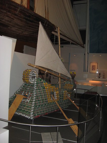 Model boat constructed from stacked aluminum cans, with sails and oars, displayed indoors
