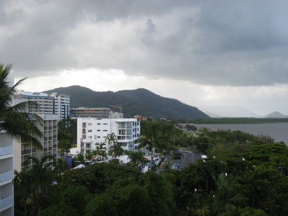 City buildings and green trees with mountains in the background under a cloudy sky