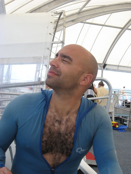 Man with bald head and hairy chest wearing a blue rash guard, eyes closed, on a boat with white canopy