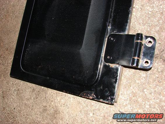 Close-up of a black vehicle door with a metal hinge attached. Some rust is visible on the edges