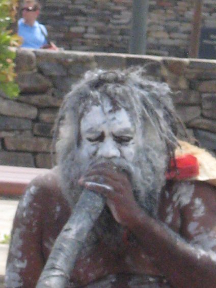 Person with white face paint and dreadlocks playing a didgeridoo