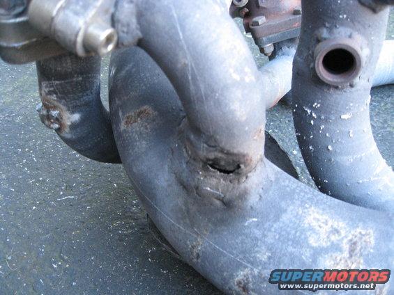Close-up of a damaged exhaust manifold with a hole in it. Supermotors.net logo visible