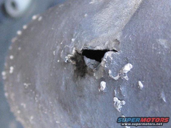 close-up of a damaged metal part with a hole and peeling paint, supermotors.net logo visible