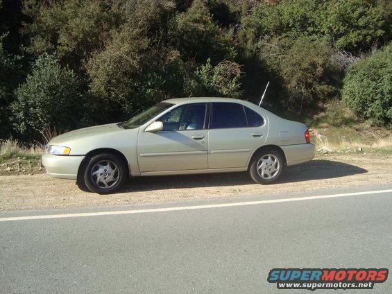 tan sedan parked on dirt shoulder next to a road, with trees and a hill in the background