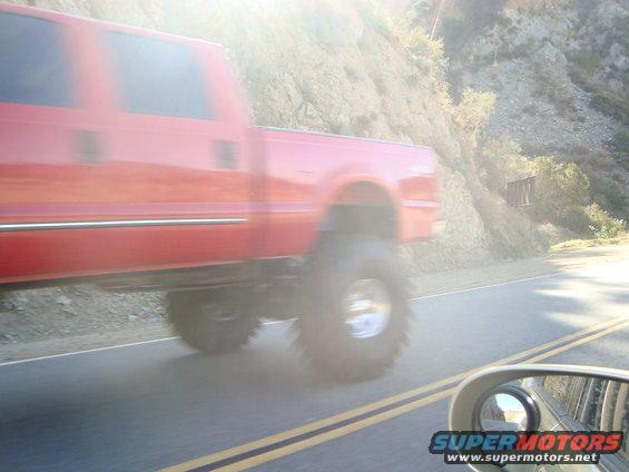 Red pickup truck with large tires driving on a road next to a hill