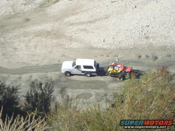 White SUV towing a trailer with two ATVs on it down a dirt road