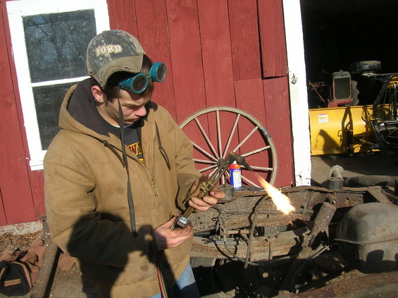guy in a Ford hat and goggles cutting metal with a torch, red barn behind him