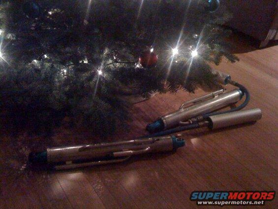 Two silver fuel pump assemblies with blue connectors and black hoses on a wooden floor near a Christmas tree