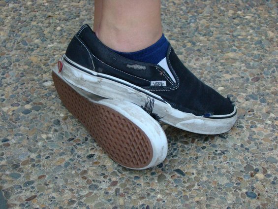 Worn black Vans slip-on shoes with blue socks on a pebbled surface