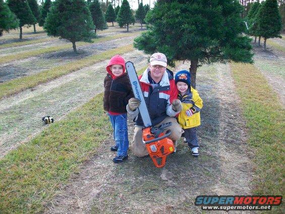 Man kneels with two children, holding a Husqvarna chainsaw in a Christmas tree farm