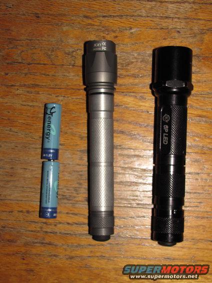 Two flashlights, one silver and one black, sit next to two AA batteries on a wooden surface