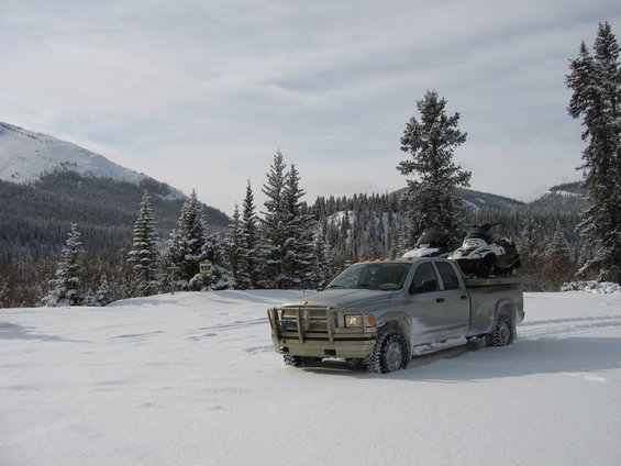 2005 Dodge 3500 pickup truck with snowmobiles in the bed, parked in a snowy landscape with pine trees and mountains