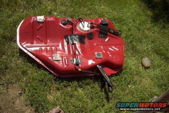 Red fuel tank with black hoses and wiring, resting on grass