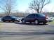 1999 Mercury Grand Marquis - Two dark sedans are parked on an asphalt surface near a body of water, with trees in the background.