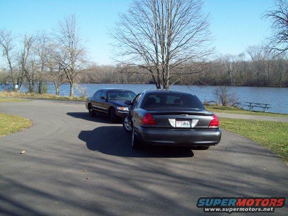 Two dark Mercury Grand Marquis sedans parked on asphalt near a lake. The rear of the closer car shows its license plate