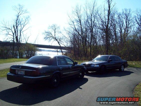 Two dark sedans parked on a paved path near a body of water, one in the foreground angled from the rear, the other behind it