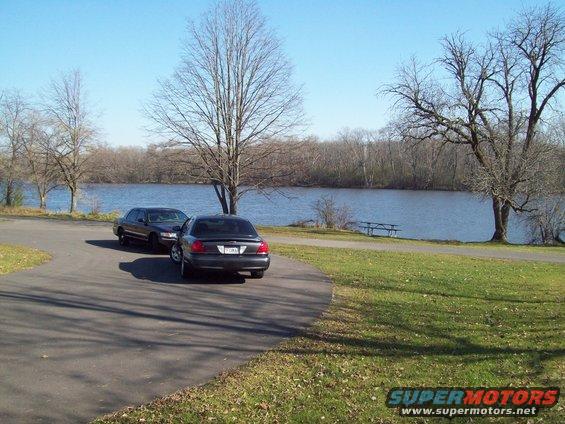 Two dark sedans parked on asphalt near a lake, one behind the other
