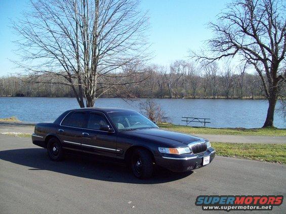Dark sedan parked by a lake, 3q-front view. Black wheels, chrome trim, tinted windows. Water and trees in background