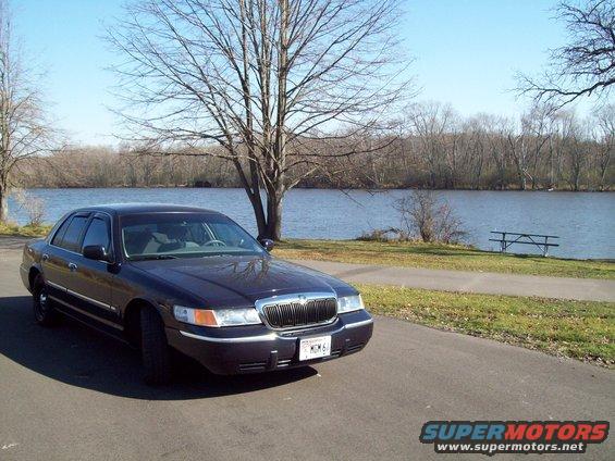 1999 Mercury Grand Marquis parked on paved path next to a lake with trees in background