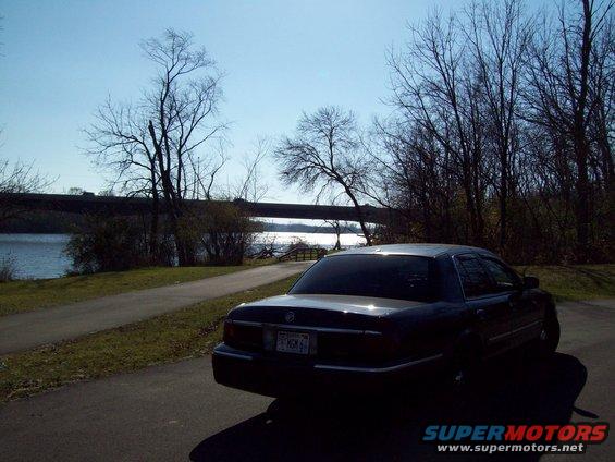 Dark sedan parked on paved path near a lake with a bridge in the background