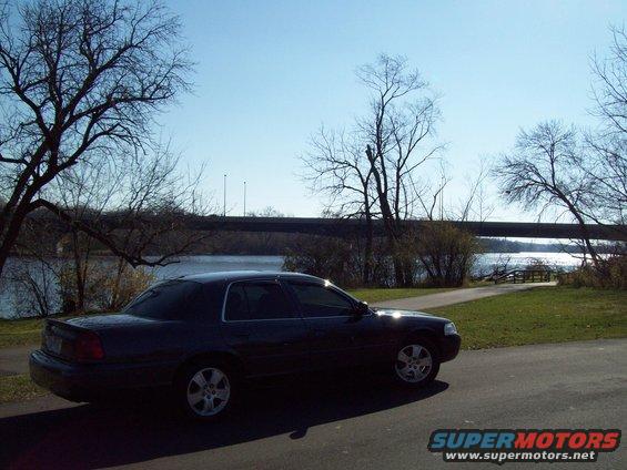 Dark sedan parked near a body of water with trees and a bridge in the background