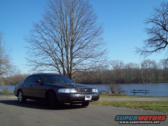 Dark sedan parked on asphalt road, facing left, with a lake and trees in the background