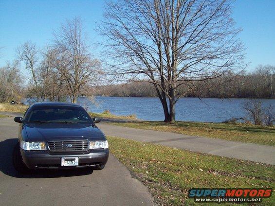 Dark sedan parked on paved path next to a lake, bare trees in background