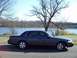1999 Mercury Grand Marquis - A dark sedan is parked on a paved surface next to a calm body of water under a bright sky.