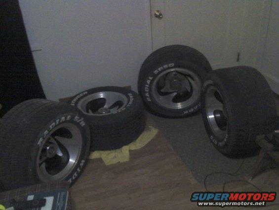 four aftermarket tires and rims stacked and leaning against a wall