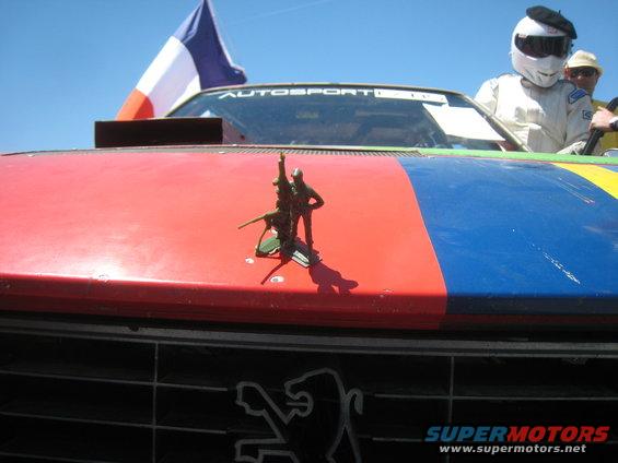 Toy soldiers hood ornament on a red, blue, and yellow striped car hood with a French flag and a Peugeot logo visible on the grille