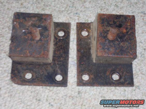 Two rusty metal mounting brackets with rubber blocks and protruding bolts, resting on a carpeted surface