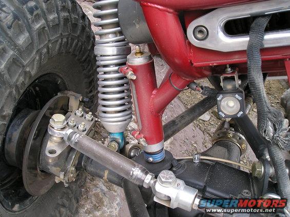 Close-up of a custom off-road vehicle's front suspension and steering linkage, showing a large tire, coilover shock, and red tubular frame