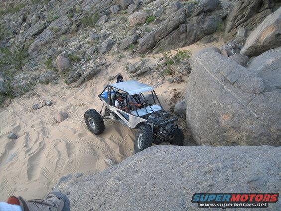 Off-road buggy with a roll cage and exposed engine, climbing a sandy, rocky slope