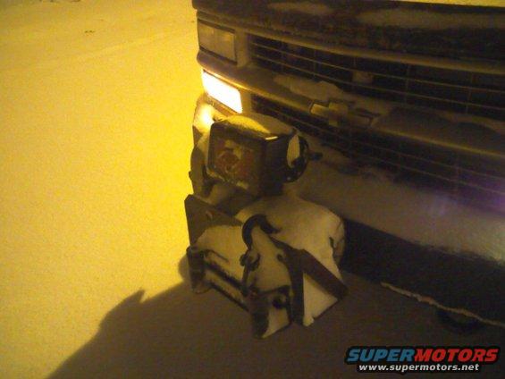Front bumper of a vehicle covered in snow, with a snow plow attachment and a yellow headlight visible