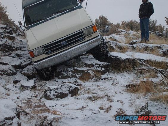 1987 Ford Bronco navigating rocky, snow-covered terrain. A person stands on a ridge in the background