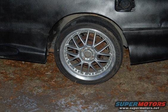 Close-up of a car's rear wheel and fender. The wheel is a multi-spoke alloy, and the fender is black with a visible fuel door