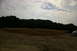 1986 Ford Bronco - A silver car is parked on a dirt lot with trees in the background under a cloudy sky.