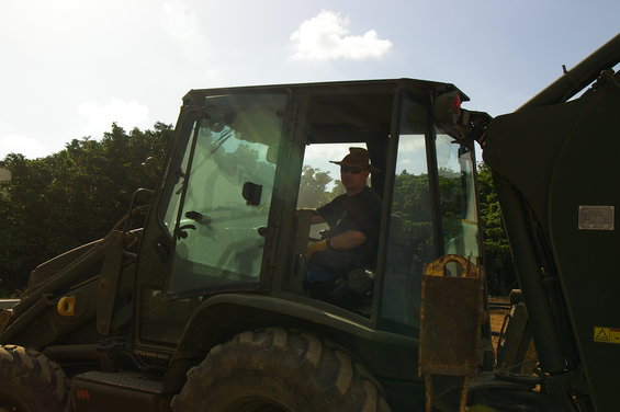Green backhoe loader with a man wearing a hat and sunglasses in the driver's seat, viewed from the side