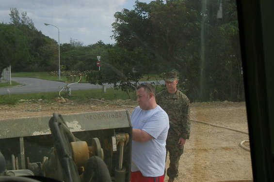 Two men, one in a t-shirt and shorts, the other in military uniform, stand near a piece of heavy equipment. Text is visible on the equipment
