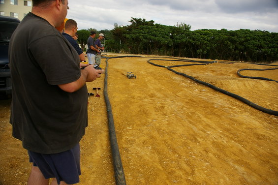 Guys controlling remote control cars on a dirt track. One car is in motion