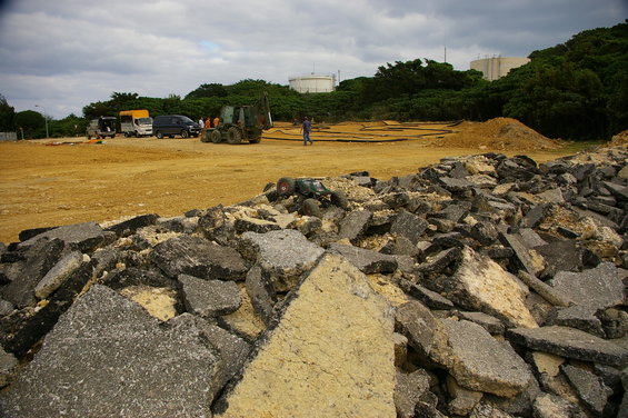 Construction site with pile of asphalt debris in foreground, trucks, a van, and a loader in background