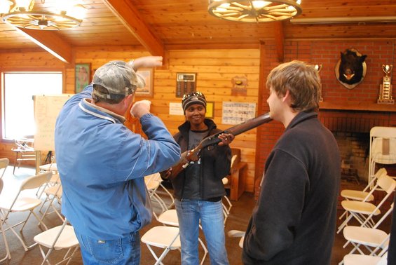 Man in blue jacket shows a rifle to two younger people in a room with wood paneling and chairs