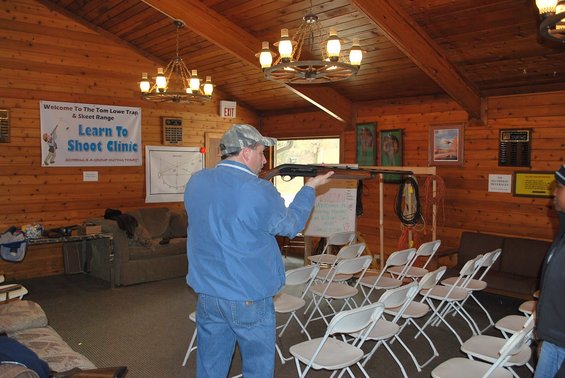 Man in blue jacket and camouflage hat holding a shotgun indoors, facing rows of chairs