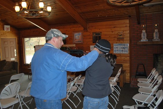 Man in blue jacket shows a woman how to hold a rifle indoors